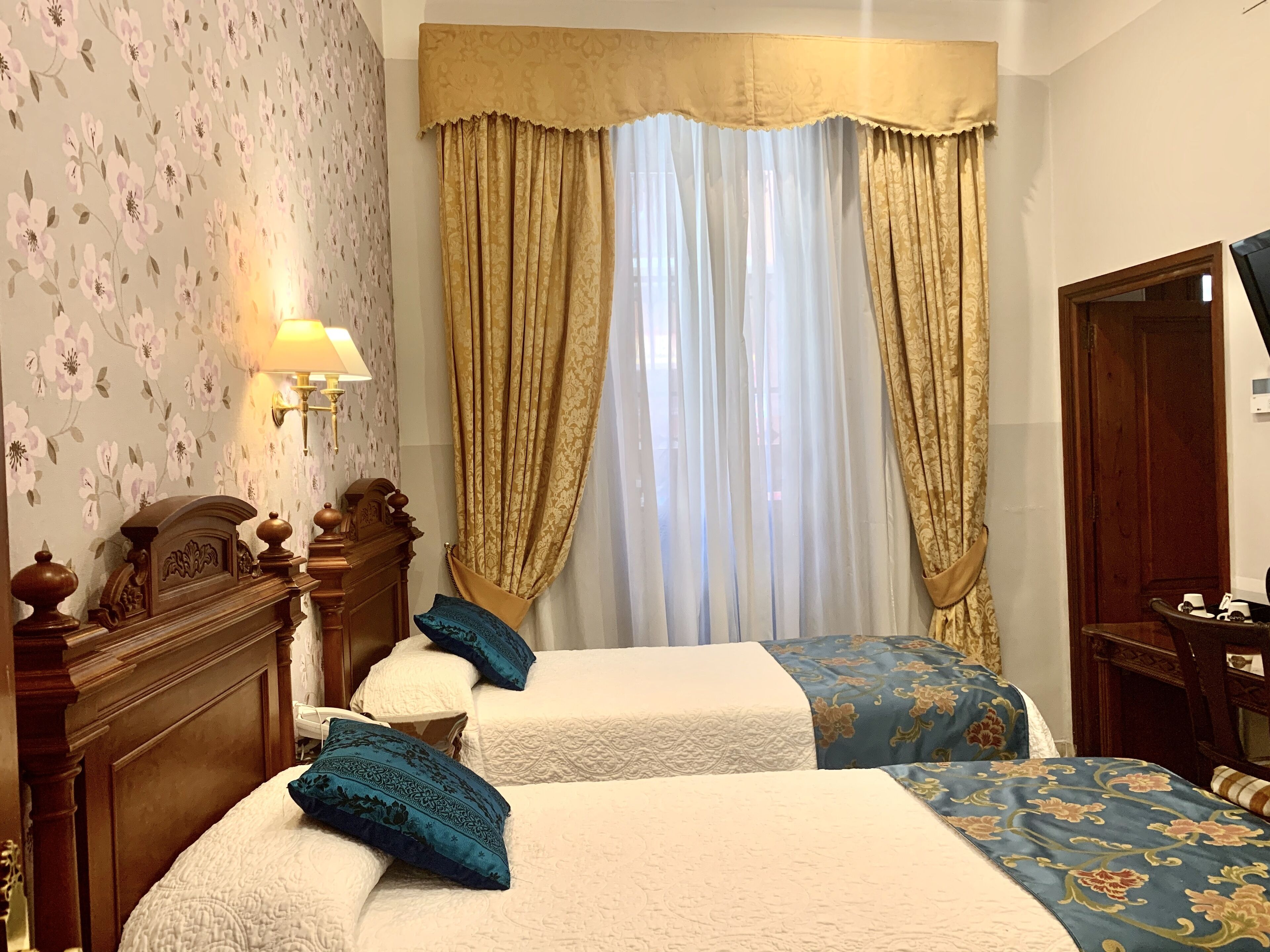 A hotel room with two beds, floral wallpaper, a wooden headboard, a large window with curtains, and a mirror.