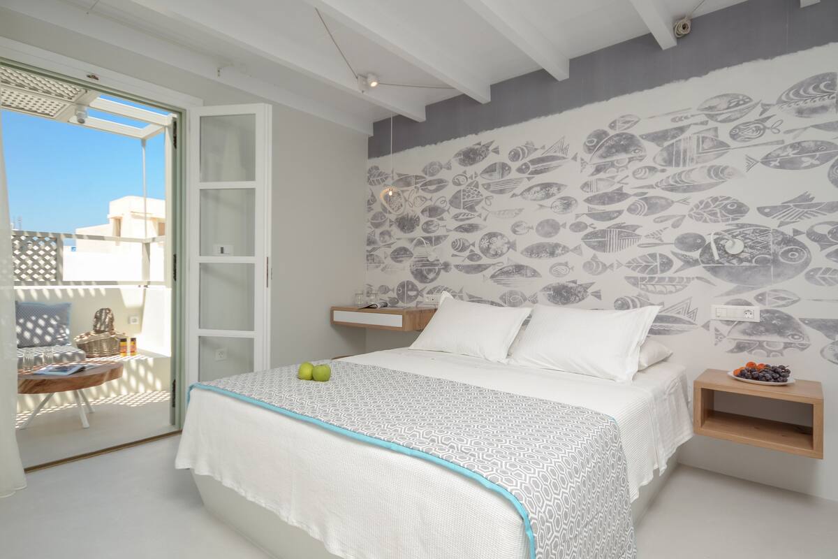 A bedroom with a bed, a wall mural of fish, a small table with a chair, and a balcony with a view of buildings.