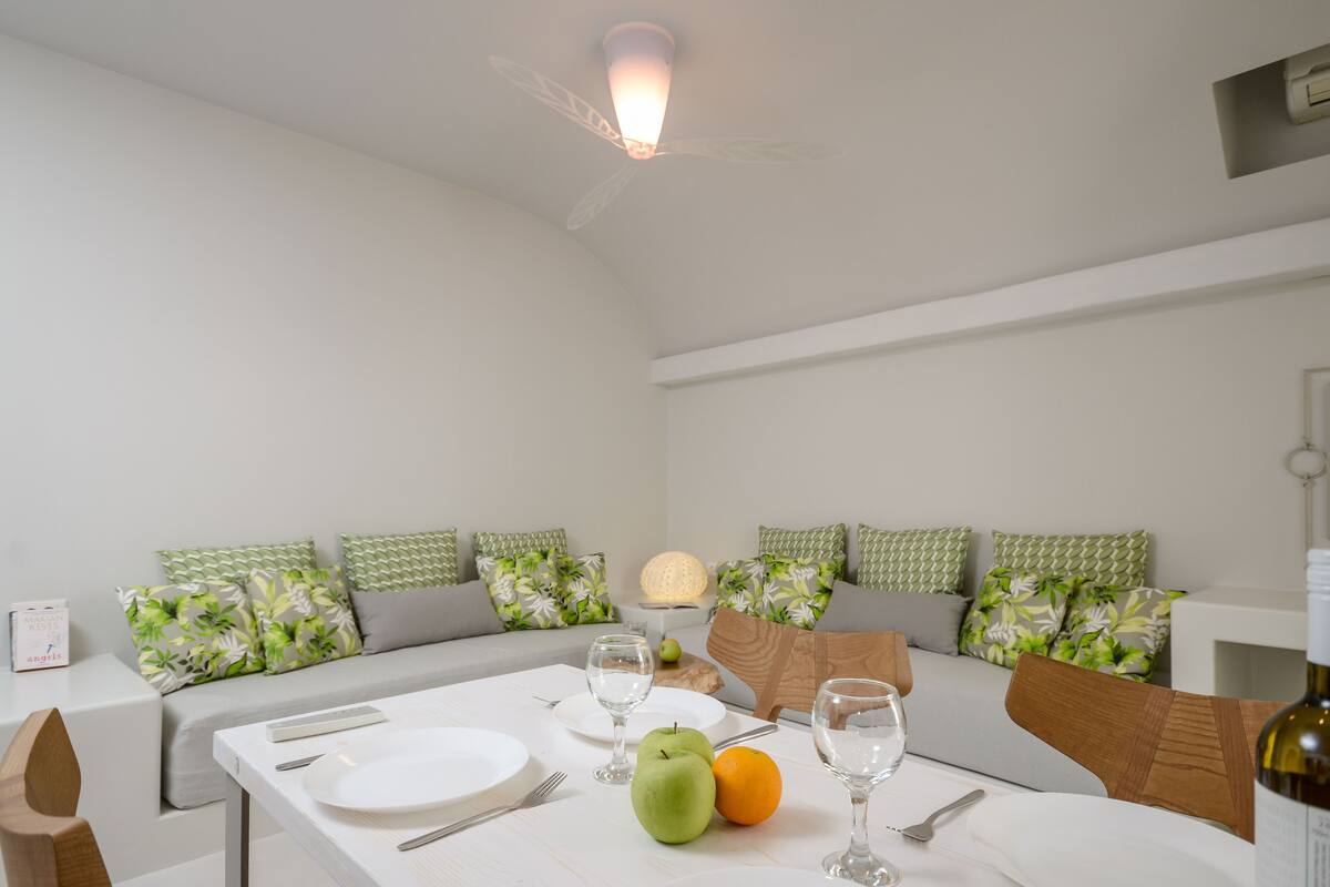A dining area with a table set for two, a sofa with patterned cushions, a lamp, and a ceiling fan.