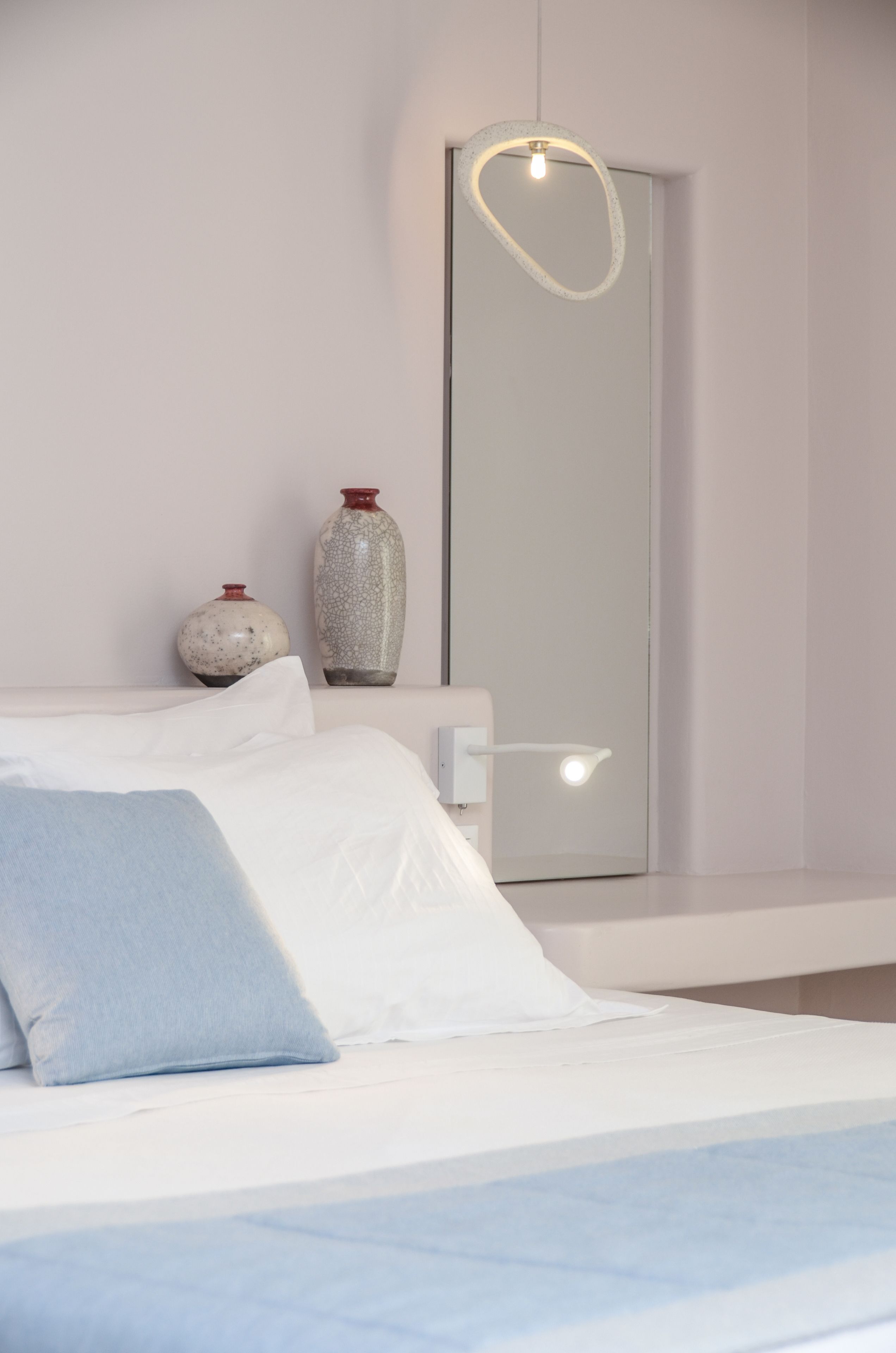 Un dormitorio con una cama, almohadas, una mesita de noche, un espejo y dos jarrones.