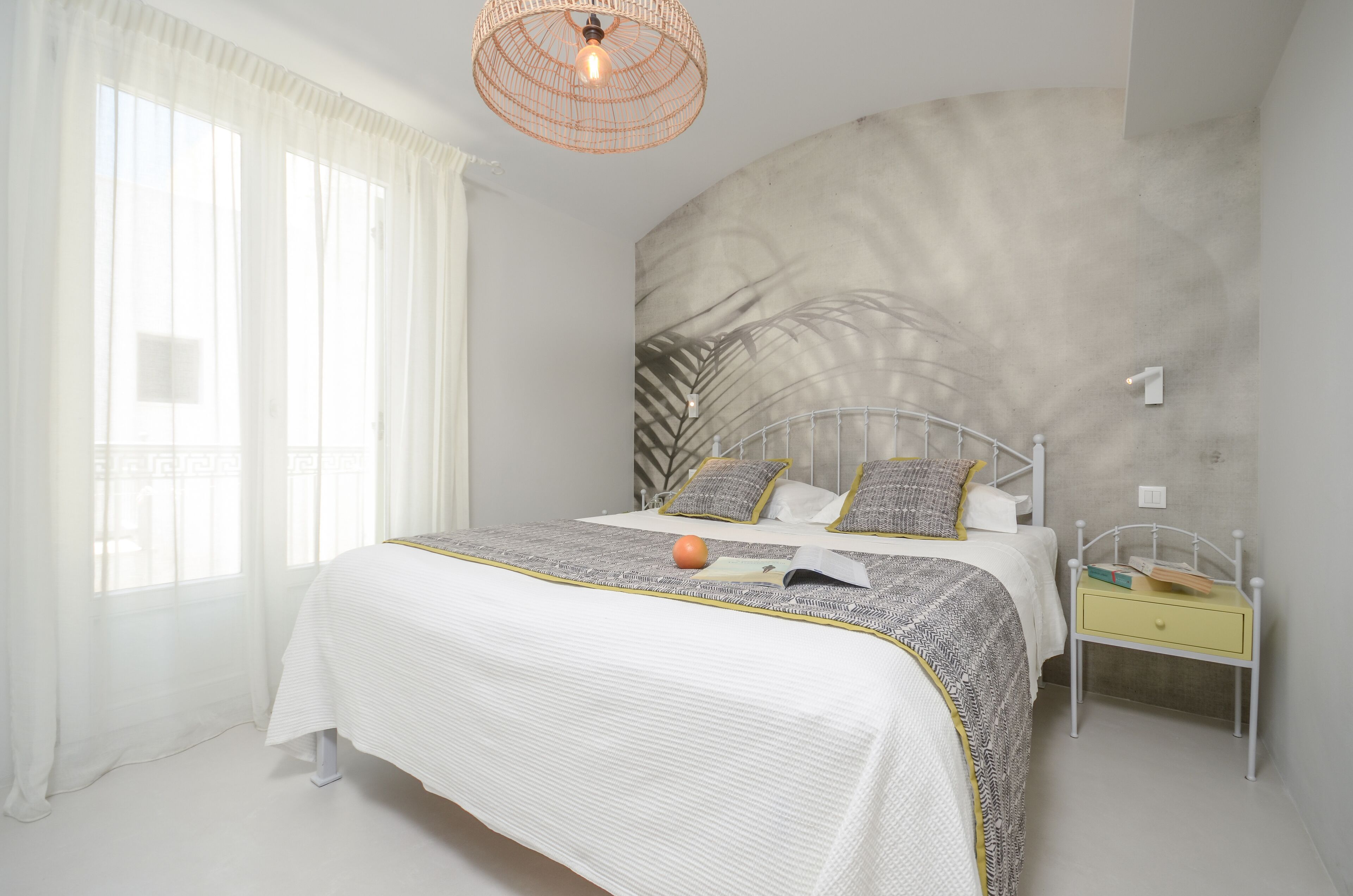 Une chambre à coucher avec un grand lit, une table de chevet, un luminaire suspendu et une fenêtre ornée de rideaux légers.