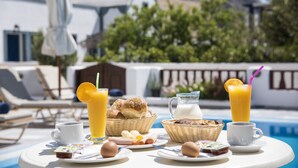 A breakfast setup with a pool view, including a pitcher of milk, orange juice, bread, and eggs. - Villa Voula (Santorini)