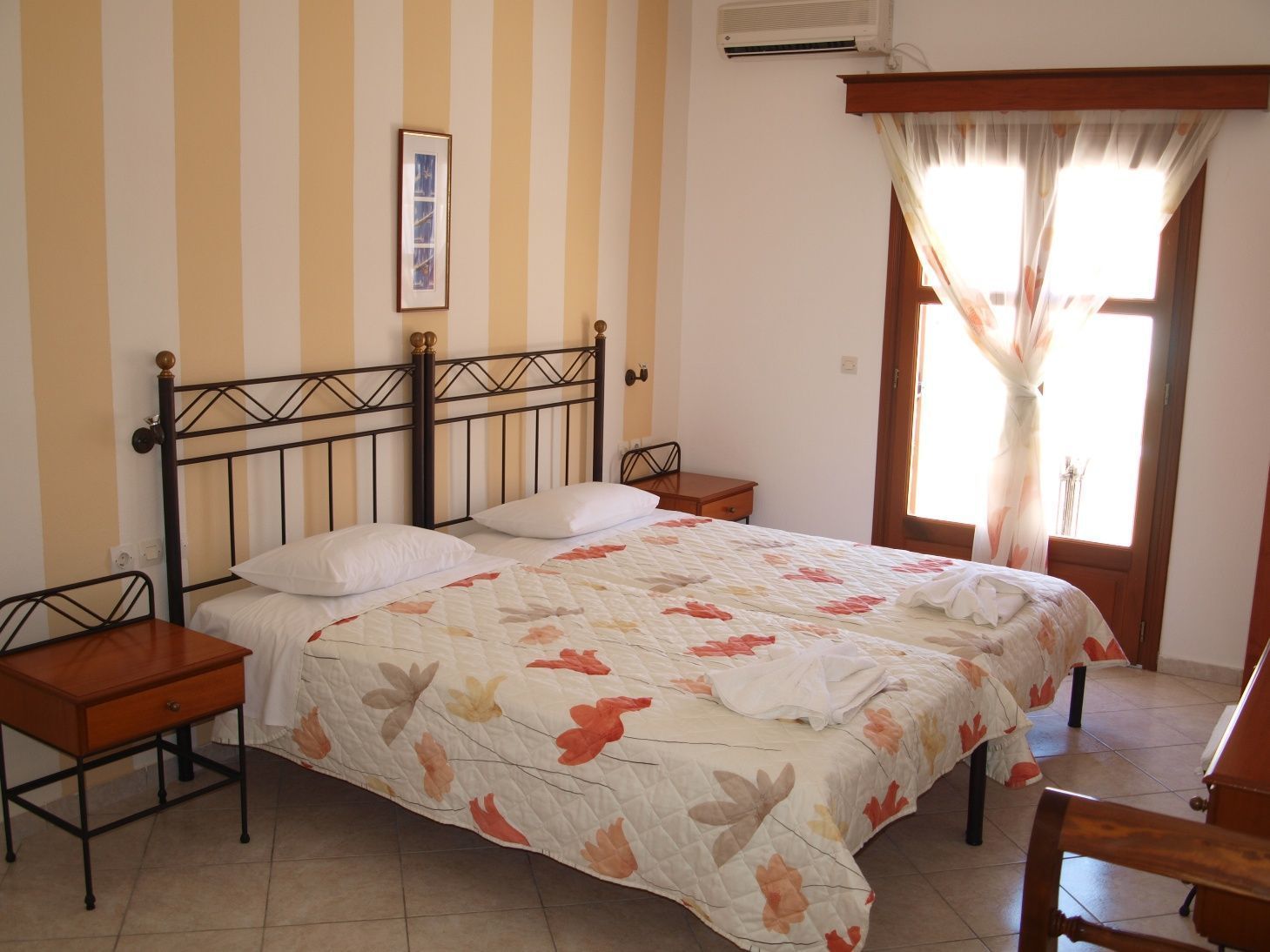 A bedroom with a bed, two bedside tables, a nightstand, a window with curtains, and a wall-mounted air conditioner.