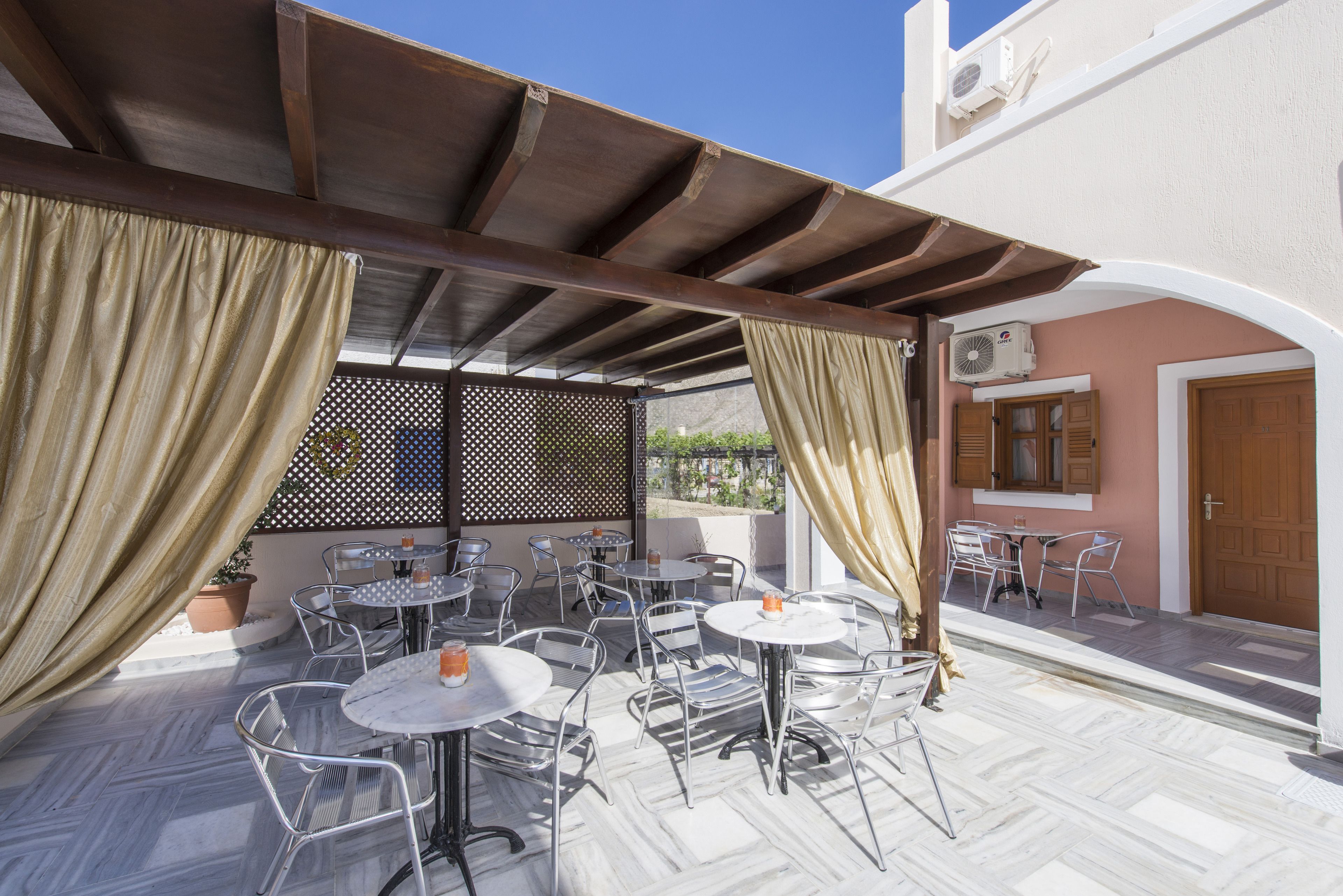An outdoor patio with multiple round tables and chairs, a pergola structure, and a view of a building with a door and window.
