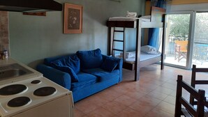 A compact living space with a kitchenette, a blue sofa, a bunk bed, and a view through a glass door to an outdoor area. - O Xenonas ton Mylon (Argos-Mykines)