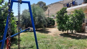 A playground with a swing set and a small wheelbarrow. There is a building with a balcony and outdoor seating. - O Xenonas ton Mylon (Argos-Mykines)