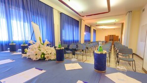 Un salón de conferencias con una larga mesa dispuesta para una reunión, cortinas azules y filas de sillas.