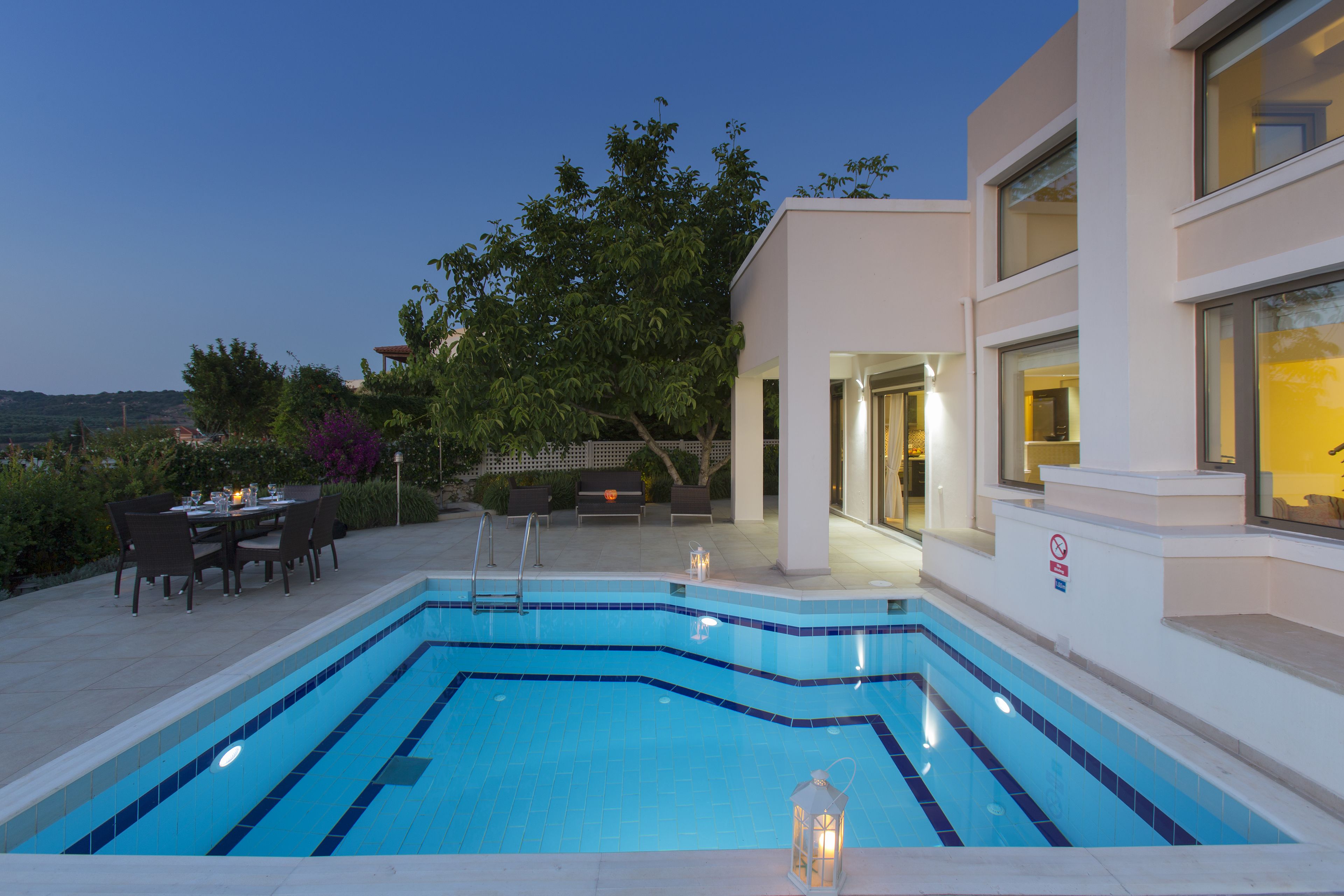 A rectangular swimming pool with blue tiles, surrounded by a patio area with outdoor furniture and a dining table.