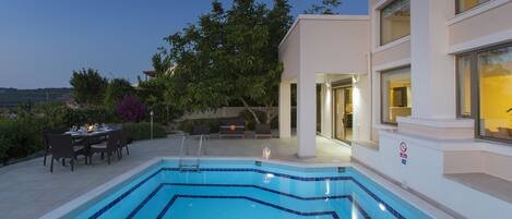 A rectangular swimming pool with blue tiles, surrounded by a patio area with outdoor furniture and a dining table.