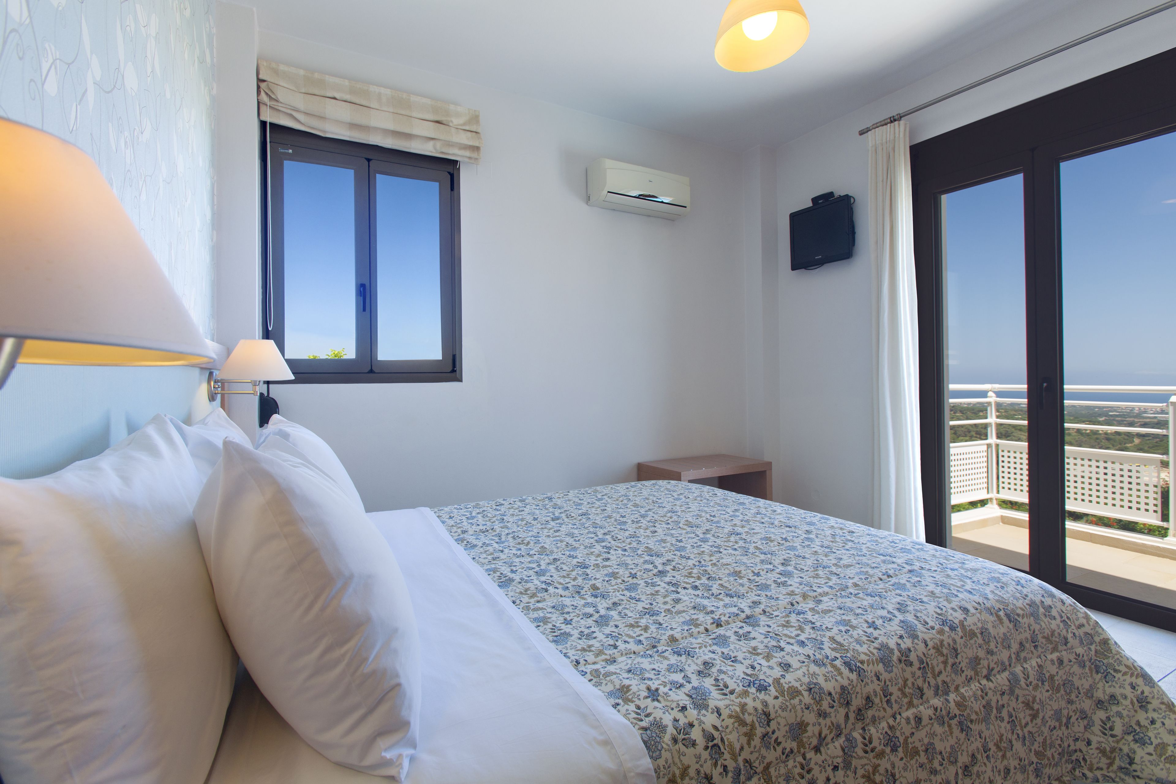 A bedroom with a bed, a nightstand, a lamp, a window, and an air conditioning unit.