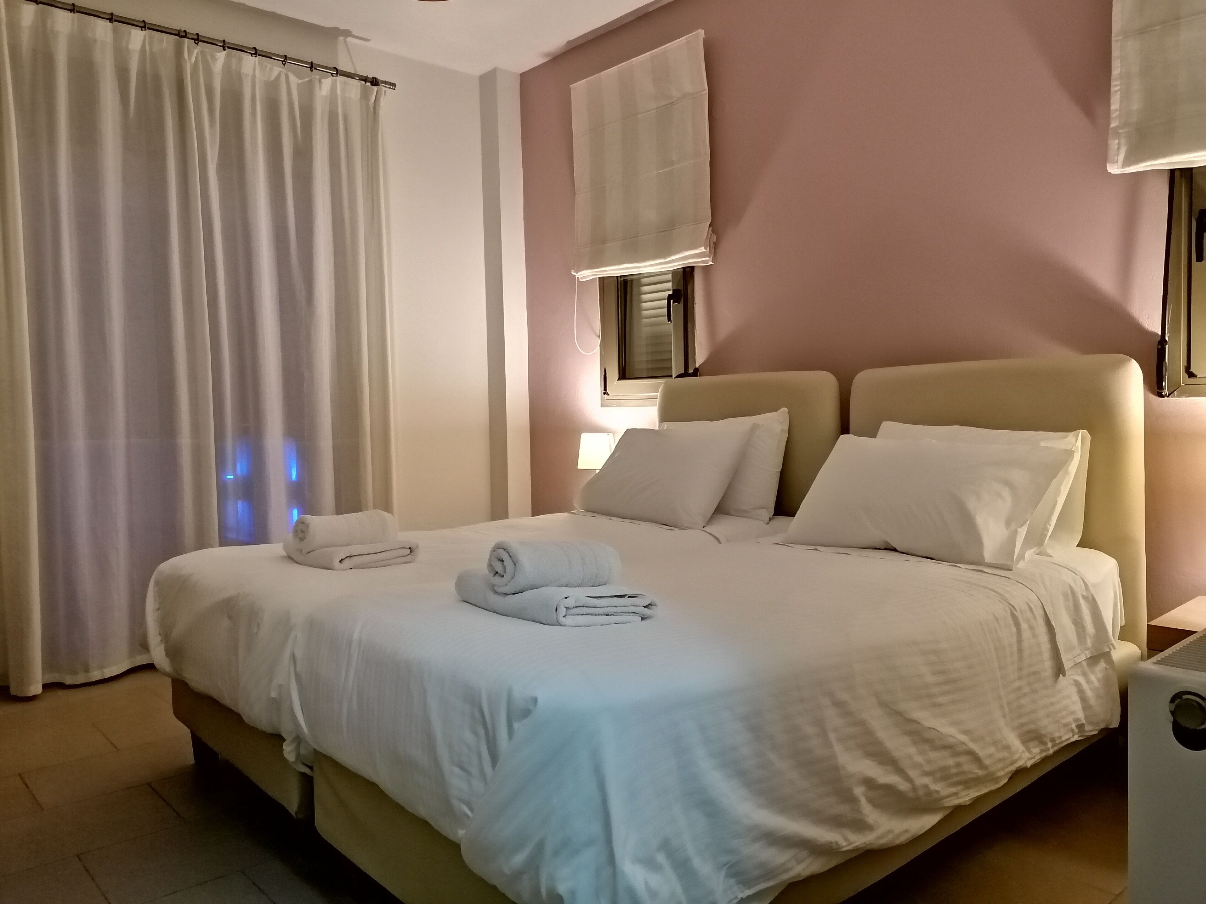 A neatly made bed with white linens and pillows, a bedside table with a lamp, and a window with a view of the outside.