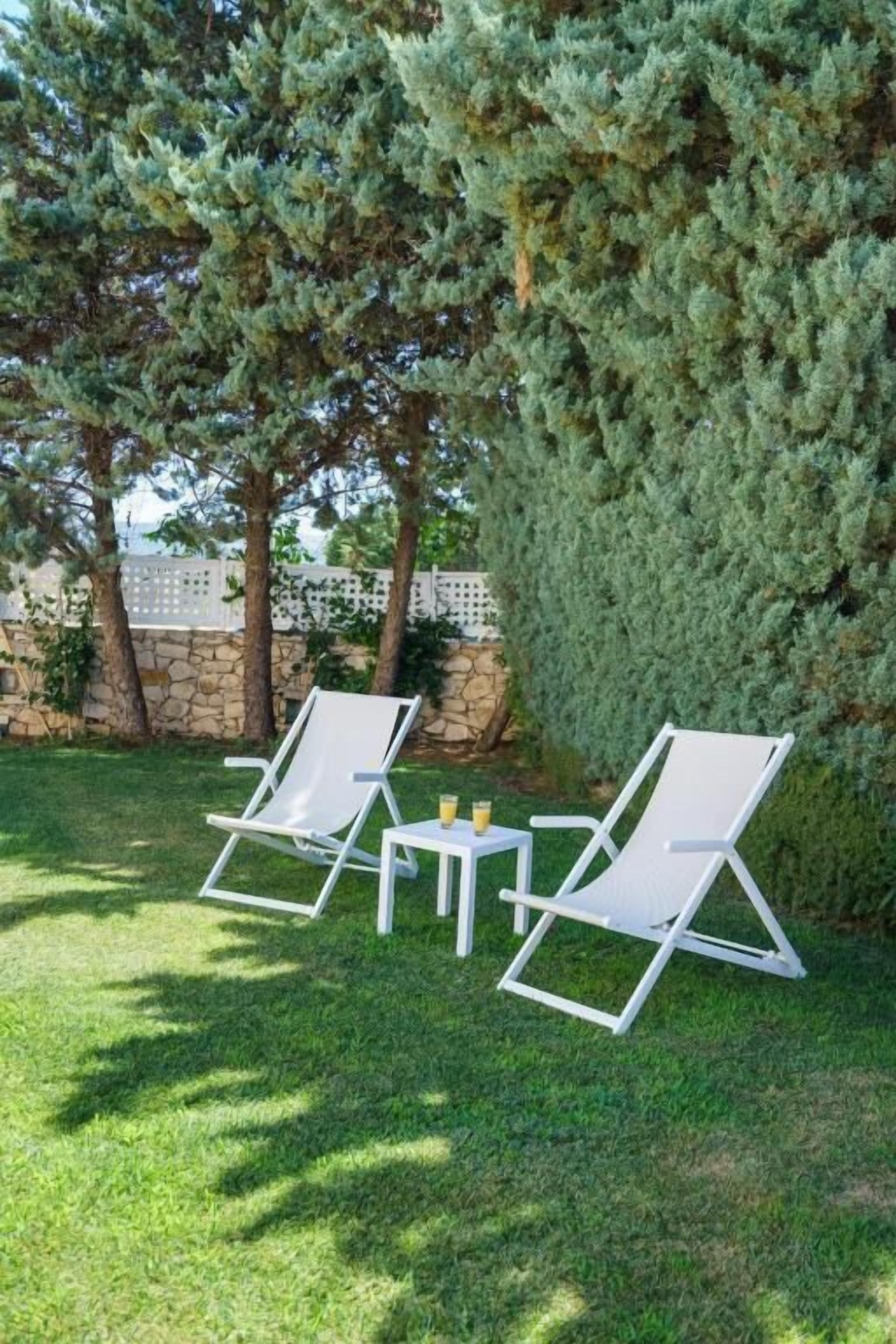 Two white lounge chairs and a small table with drinks in a grassy outdoor area, surrounded by trees and a stone wall.