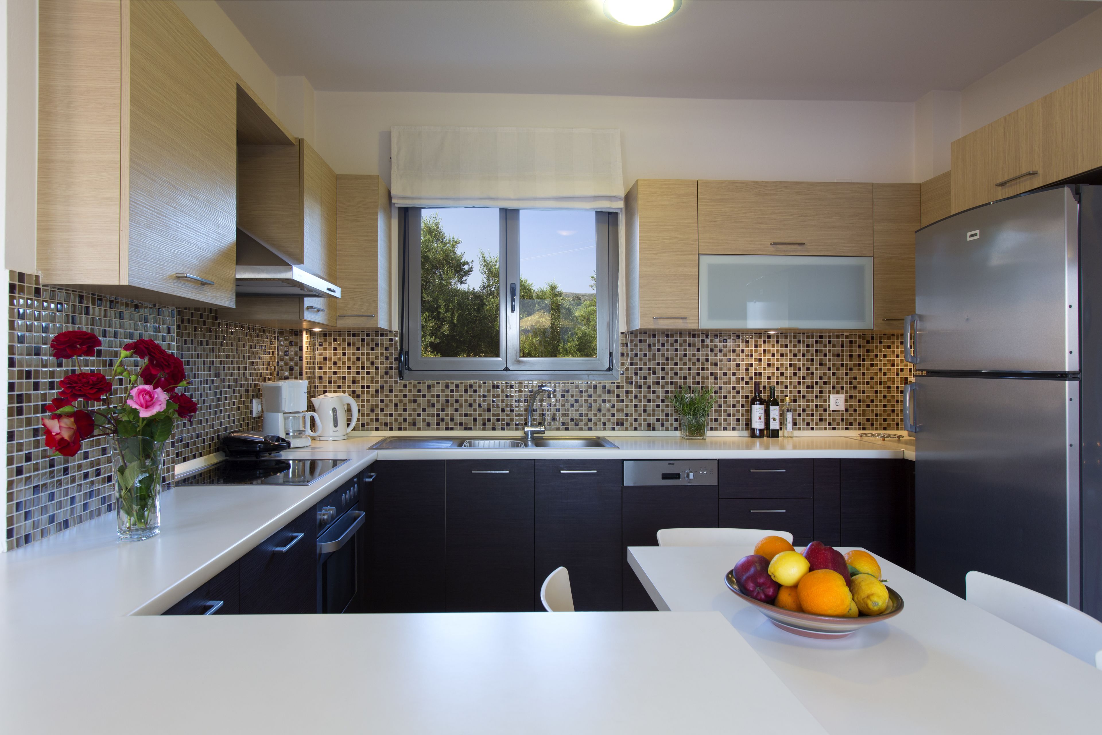 A modern kitchen with a large window, a stainless steel refrigerator, and a bowl of fruit on the counter.