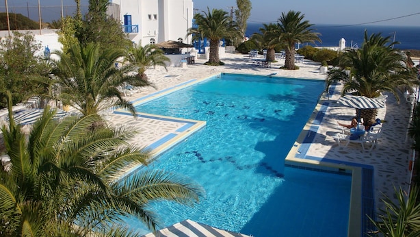 A clear blue swimming pool with a sandy deck, surrounded by palm trees and a white building with blue accents. - Hotel Alkyon (Syros)