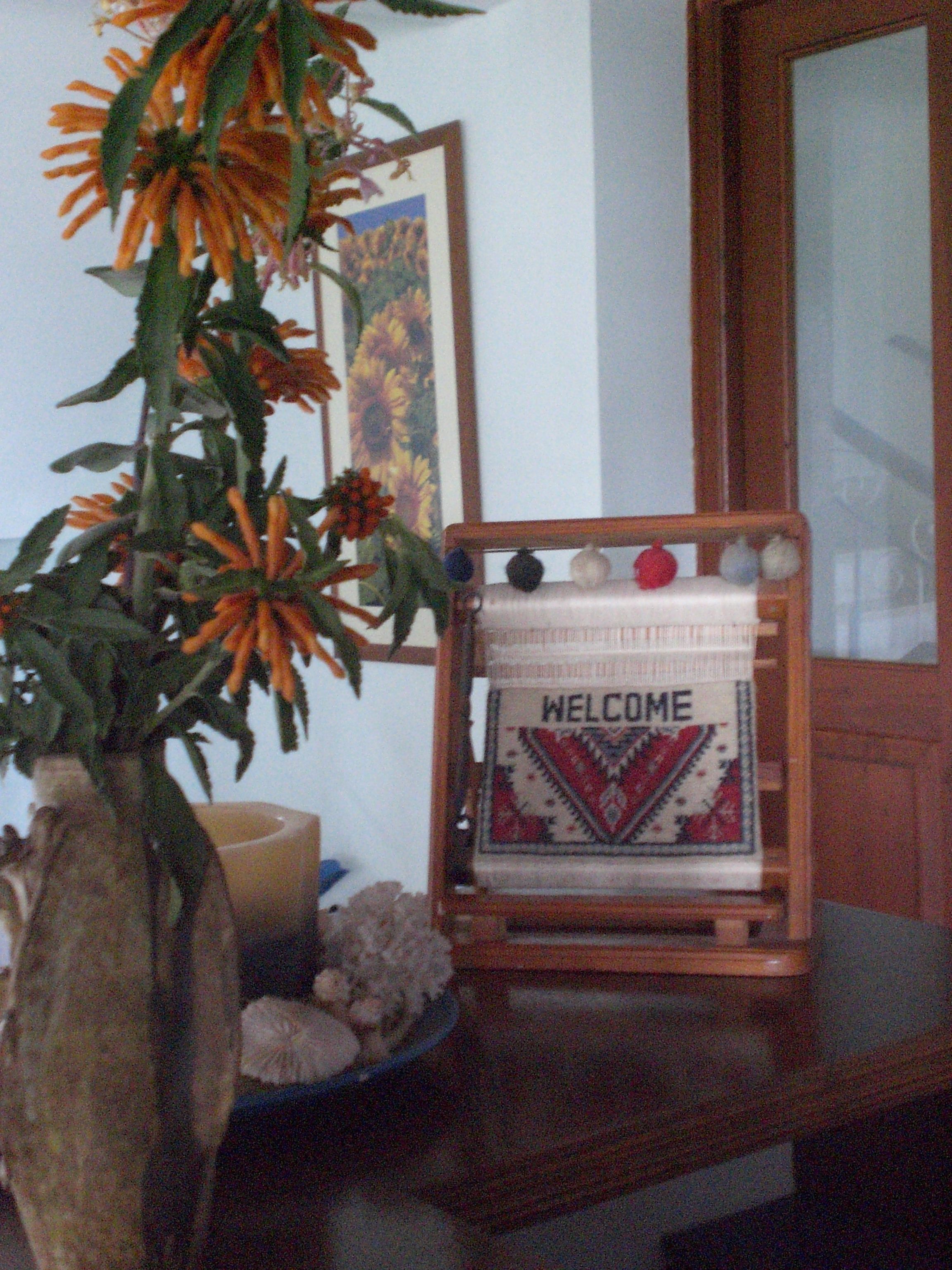 Een kamer met een houten tafel waarop een welkomstbord staat, een vaas met oranje bloemen en een ingelijst beeld aan de muur.