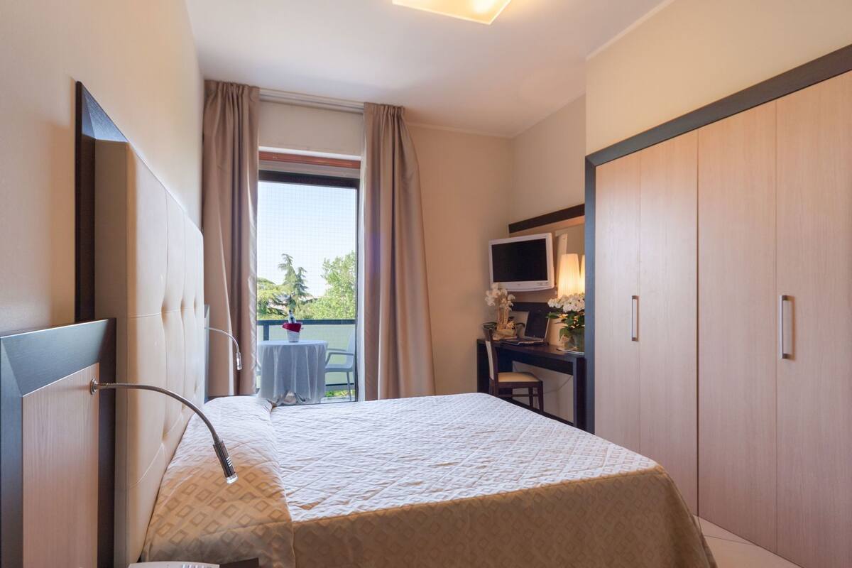 A hotel room with a bed, a desk with a computer, a TV, and a balcony with a table and chairs.