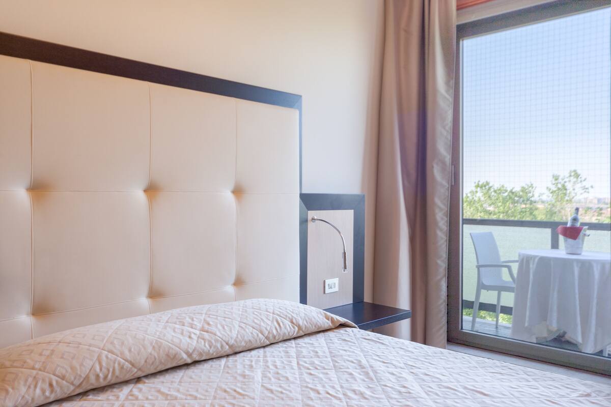 A hotel room with a large bed, a headboard, a small table with a chair, and a view of the outdoors.