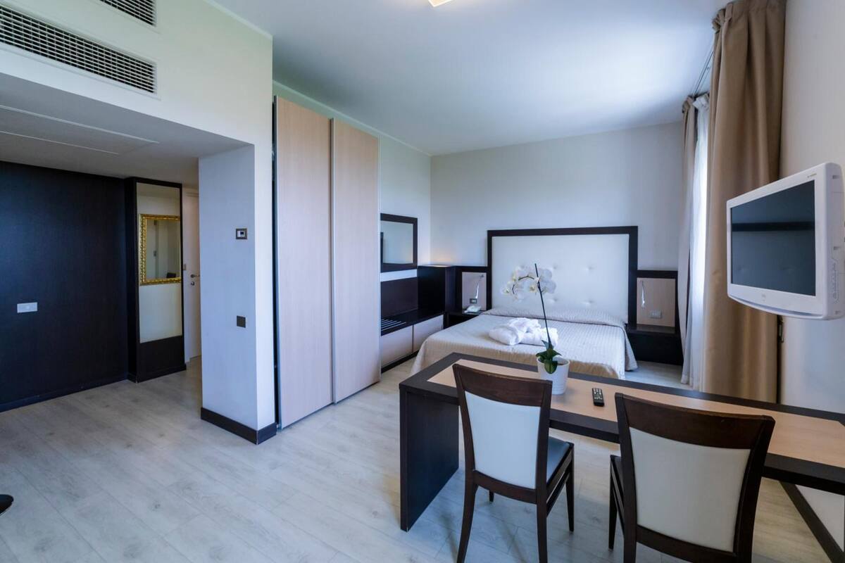 A modern hotel room with a bed, bedside table, TV, and a desk with chairs.