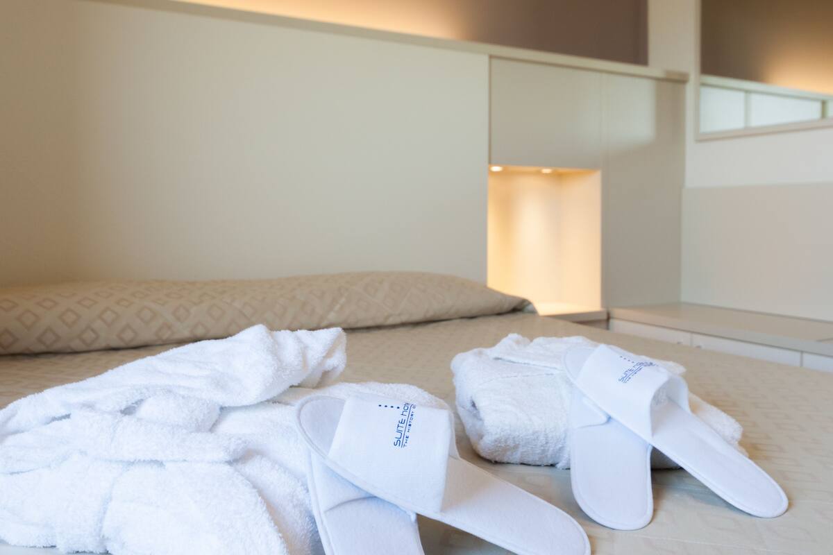 A hotel room with a neatly made bed, a pillow, and two pairs of white slippers with a logo on them.
