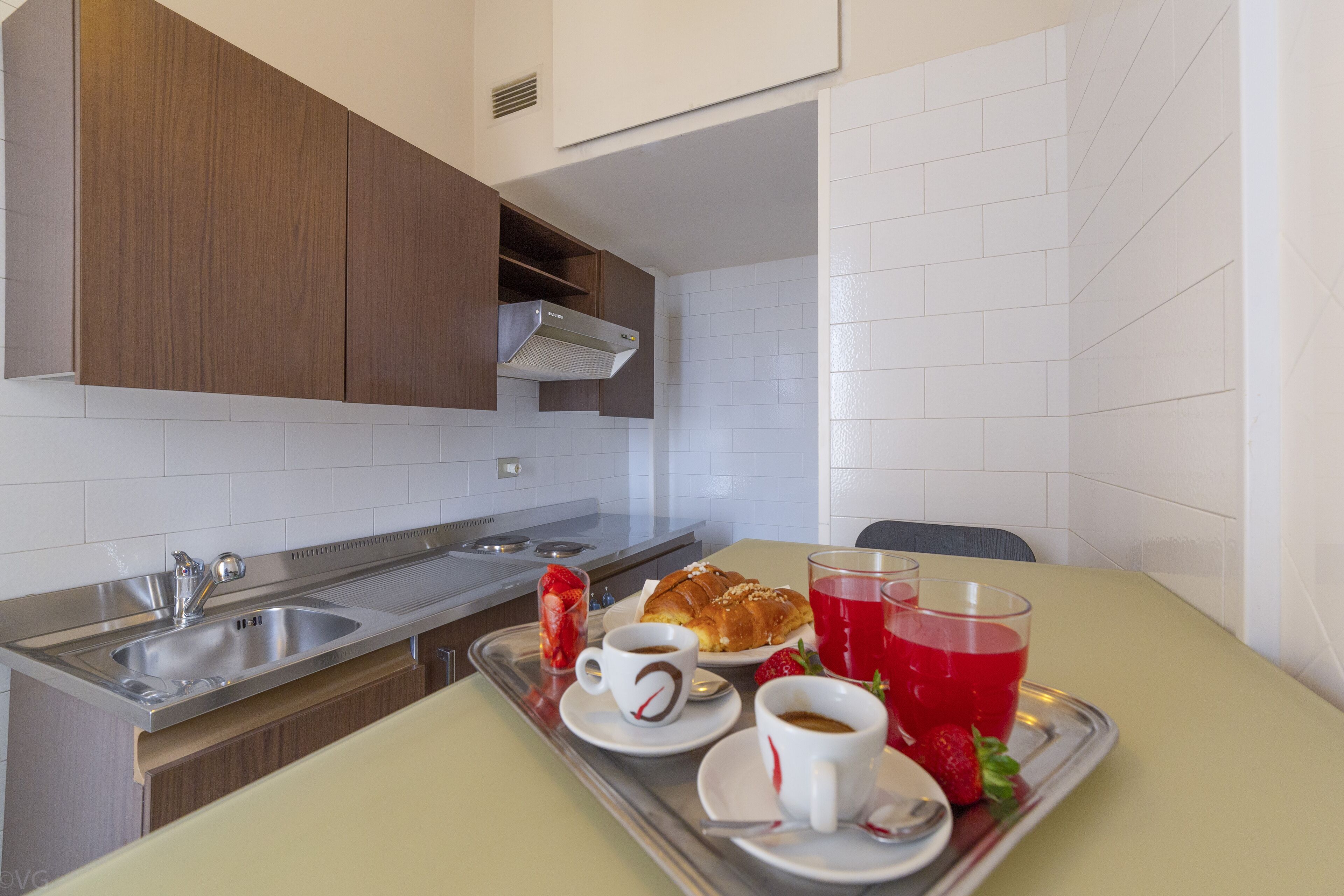 A kitchenette with a stainless steel sink, a tray with a croissant, coffee cups, and a red beverage.