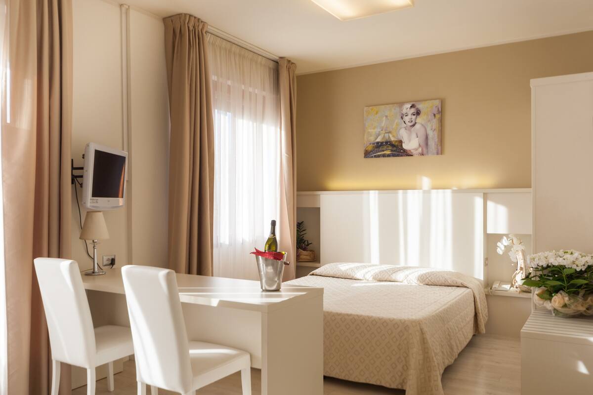 A hotel room with a bed, a desk with a chair, a TV, and a painting on the wall.