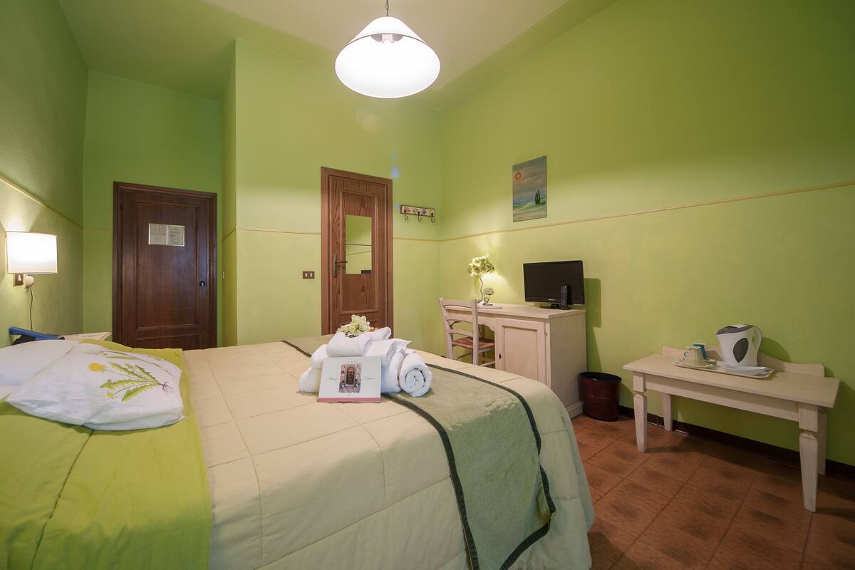 Habitación de hotel con cama, escritorio, silla, televisor y mesita con hervidor y tazas.