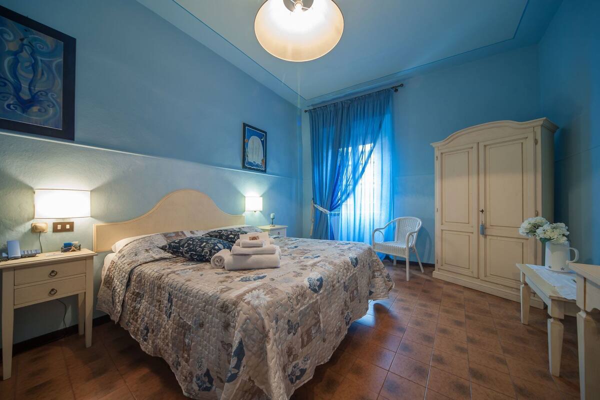 Un dormitorio con cama, mesitas de noche, una silla, armario y escritorio con lámpara.