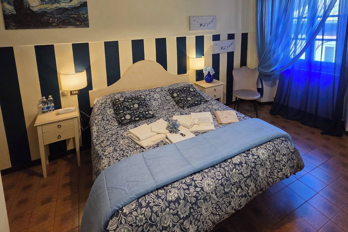 Una habitación de hotel con cama, mesitas de noche, lámpara, un cuadro y ventana con cortinas.