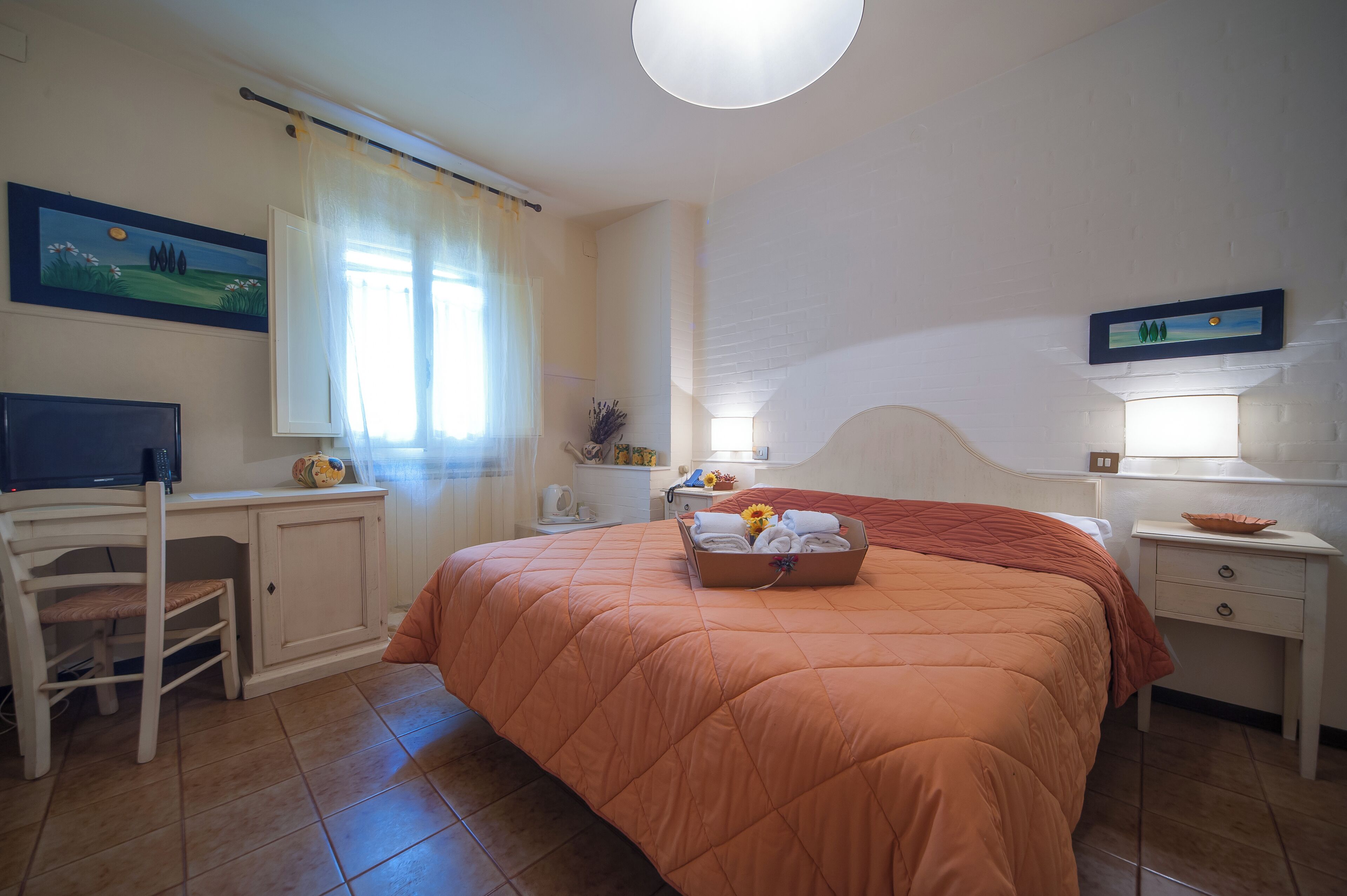 Un dormitorio con cama, escritorio, silla, mesita de noche, lámpara y un cuadro en la pared.