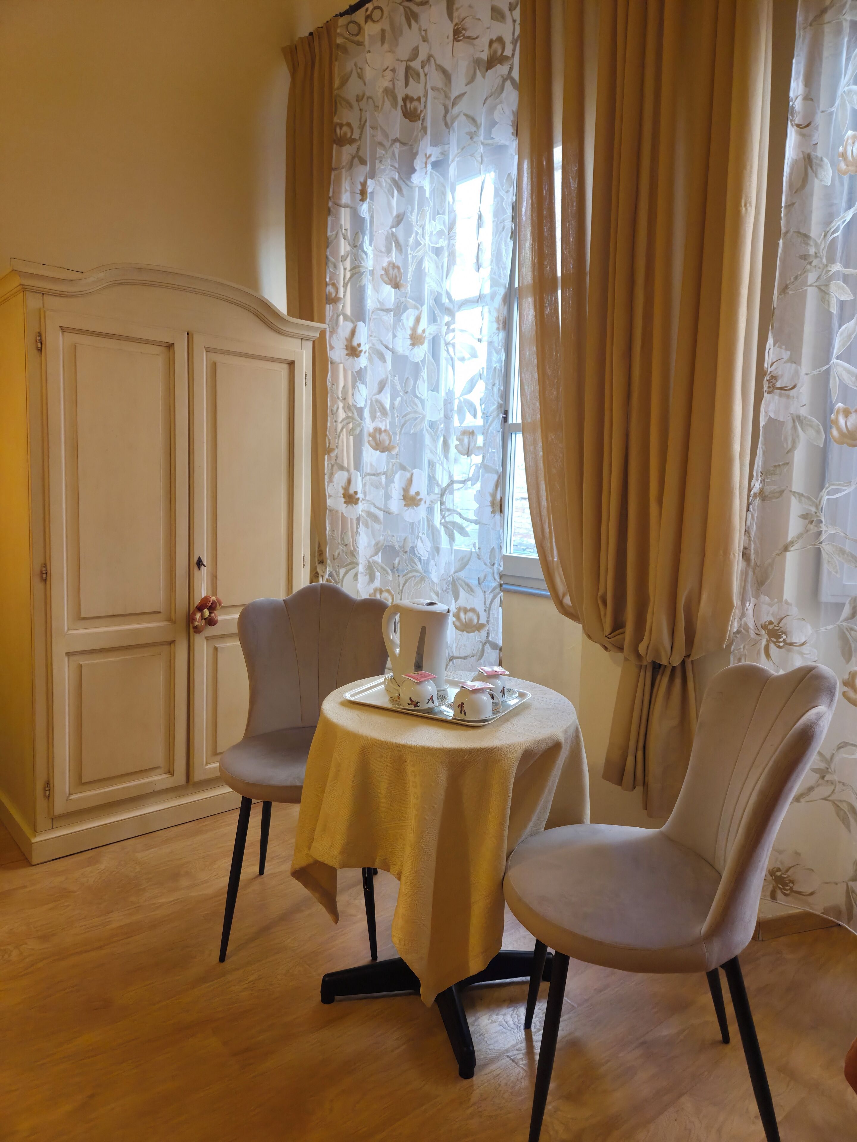 Una habitación con dos sillas alrededor de una pequeña mesa, un armario y una ventana con cortinas florales.