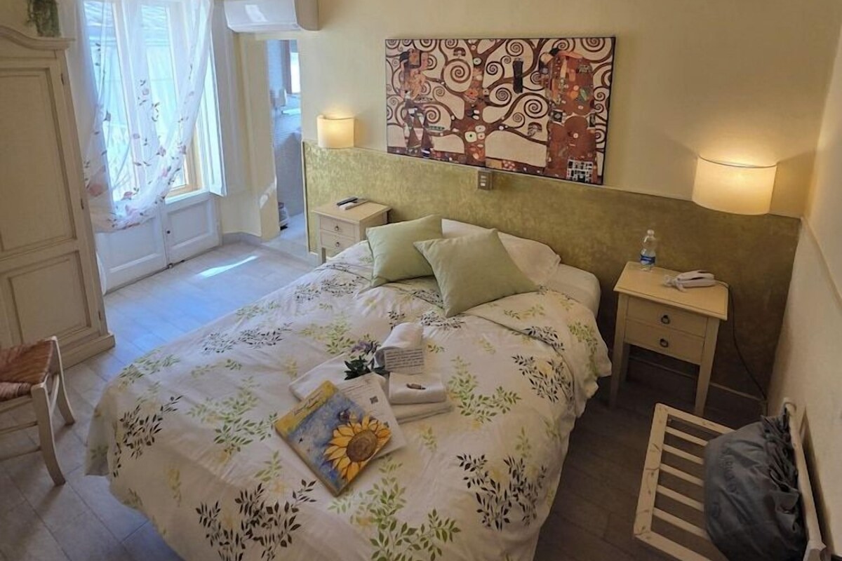 A bedroom with a bed, bedside lamps, a headboard, a window with curtains, and a wall with a painting.