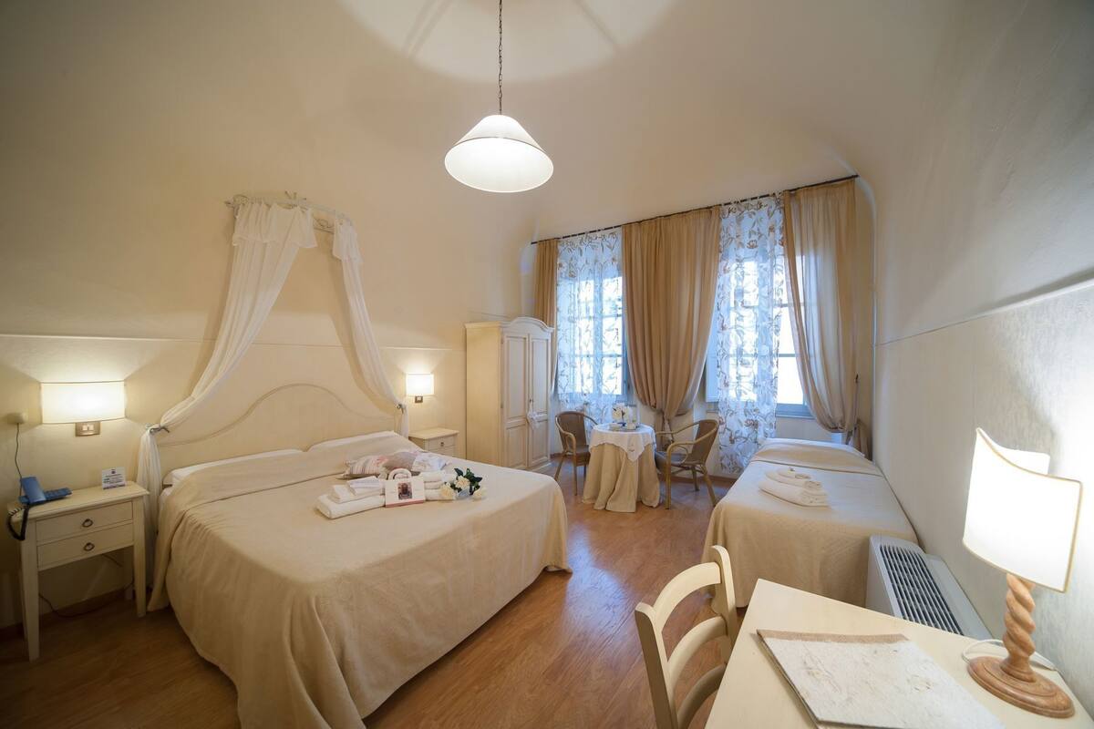 Habitación de hotel con cama, mesitas de noche, escritorio, sillas, ventana con cortinas y una lámpara colgante.