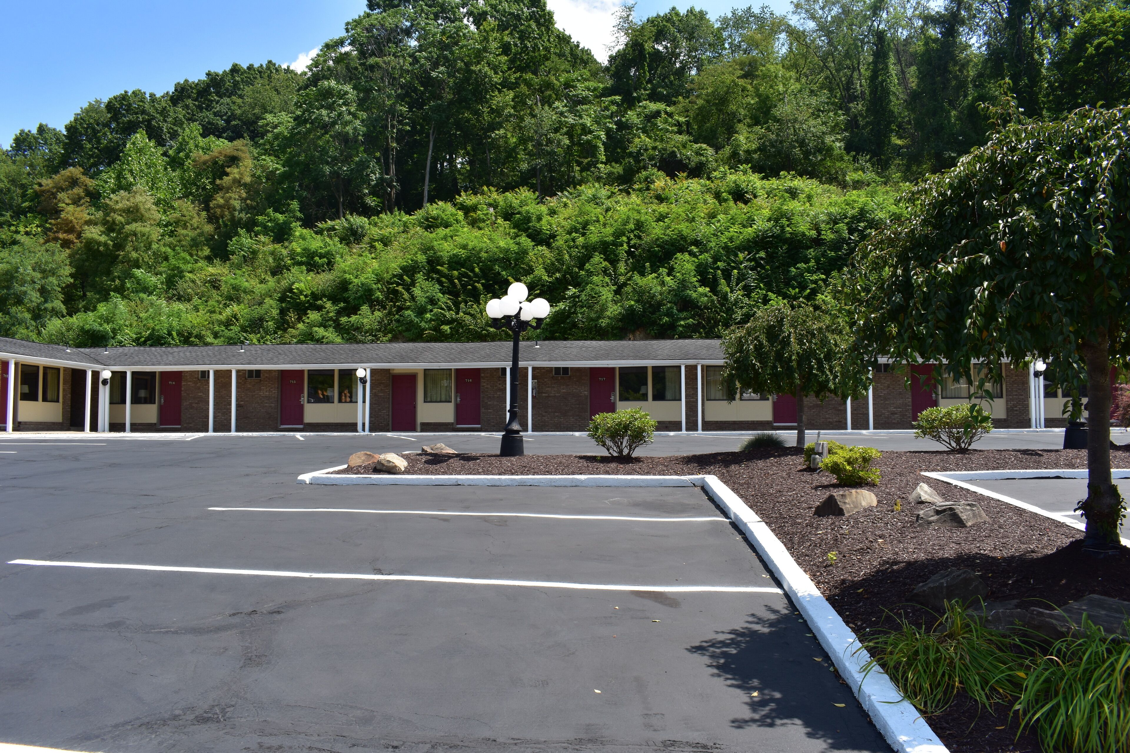 A motel with a parking lot, a small garden area with shrubs and a tree, and a building with a red and beige facade.