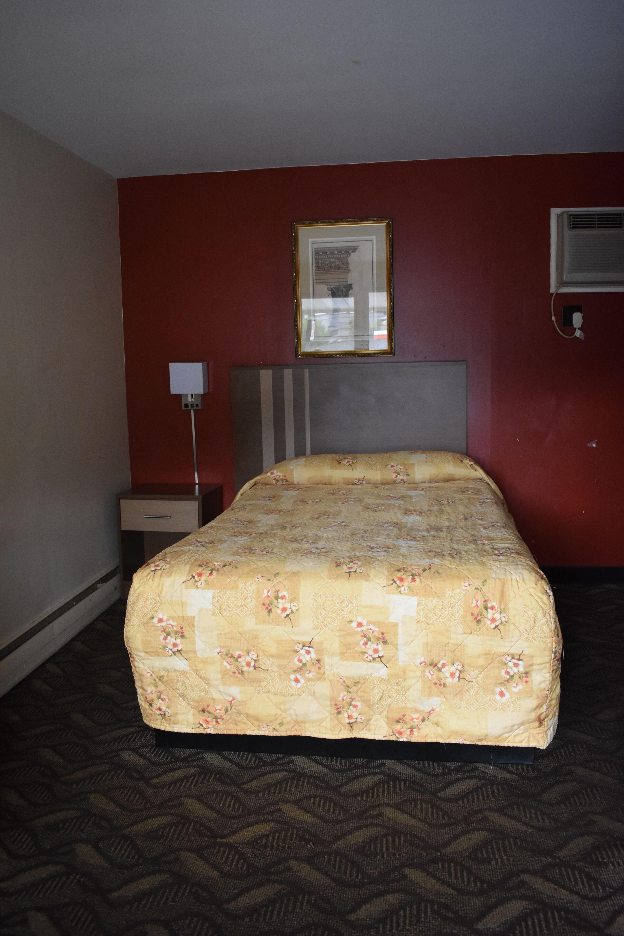 A hotel room with a bed, a nightstand, a lamp, and a wall-mounted air conditioning unit.