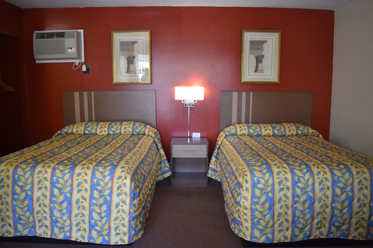 A hotel room with two beds, a nightstand, a lamp, and an air conditioner.