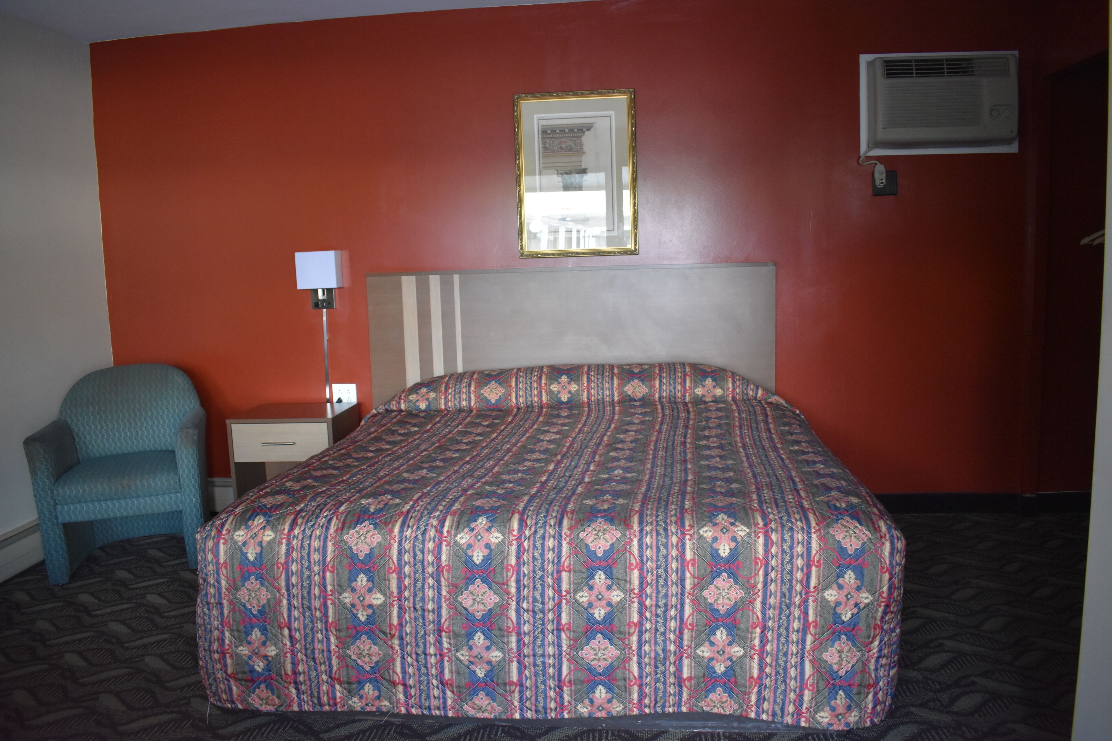 A hotel room with a patterned bedspread, a single bedside table, a chair, and a wall-mounted air conditioner.