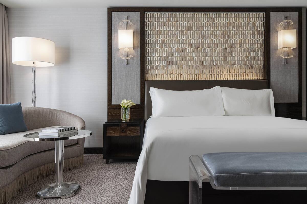 Une chambre d’hôtel avec un grand lit, une table de chevet avec une lampe, une petite table avec des livres, un canapé et un panneau décoratif au mur.