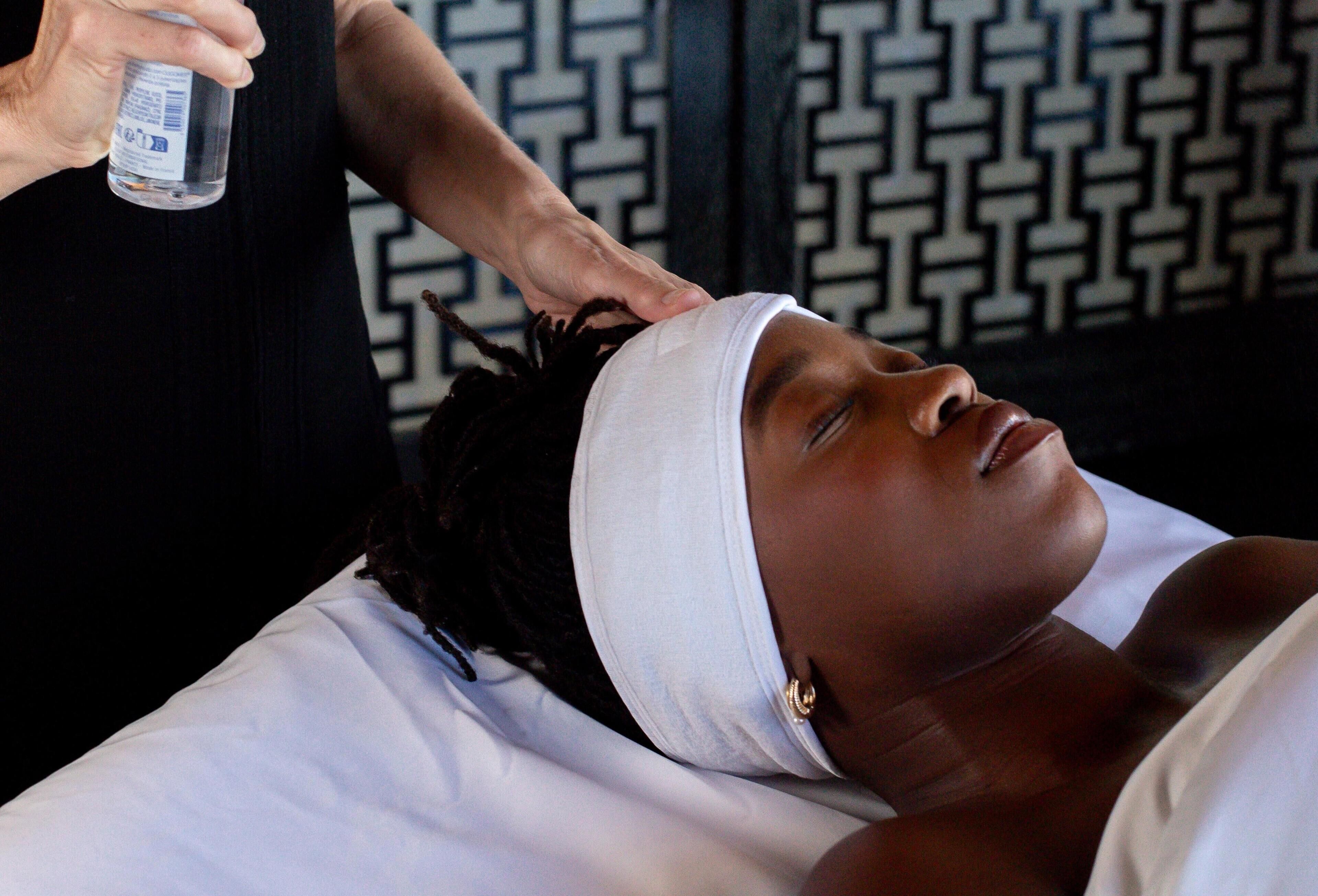 A person lying on a bed with a white pillow and headband, receiving a massage from another person.