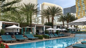 A pool area with lounge chairs and umbrellas, surrounded by palm trees and modern buildings.