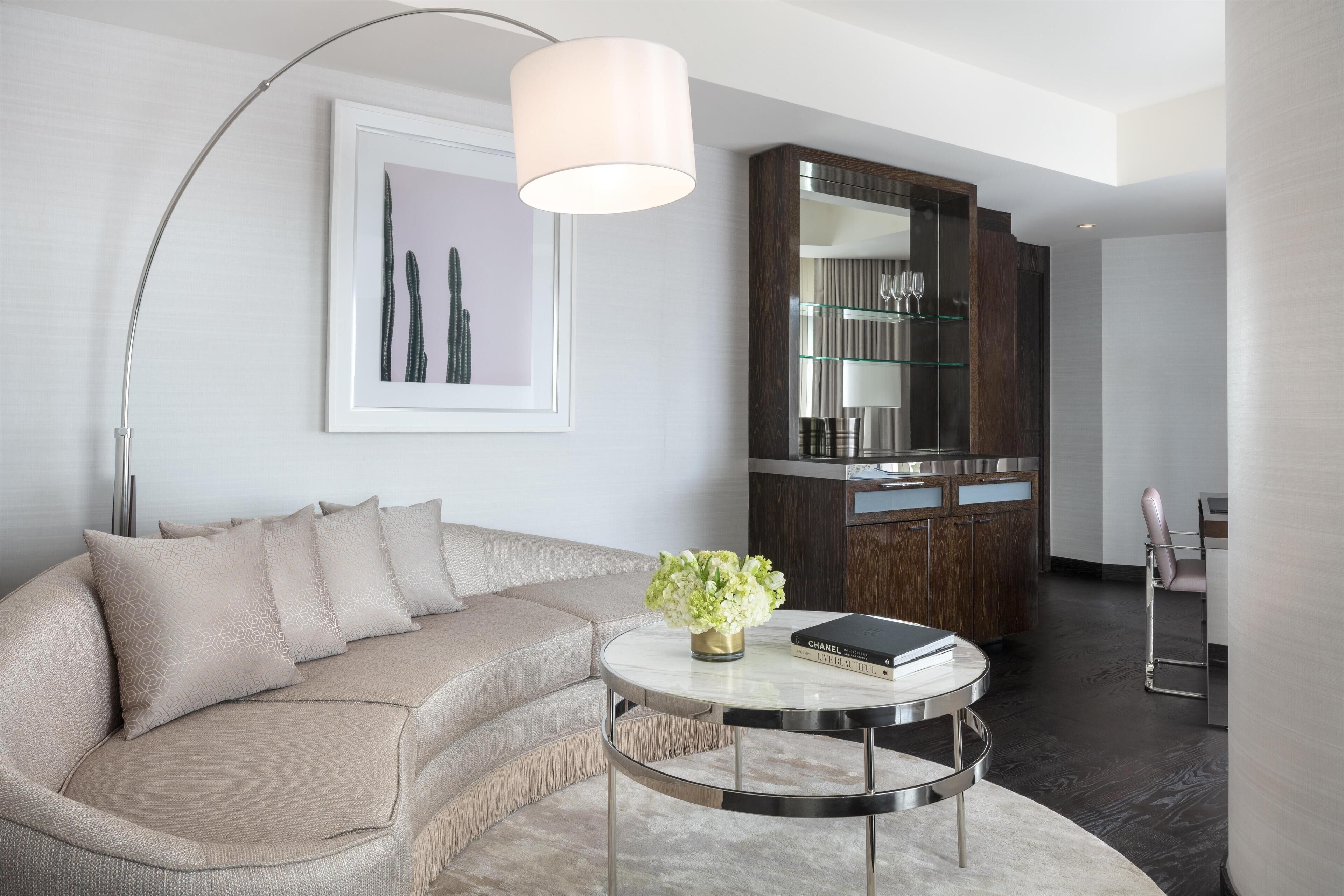 Un salon moderne avec un canapé beige, une table basse ronde sur laquelle reposent un livre et des fleurs, un grand lampadaire et un meuble avec des portes en verre.