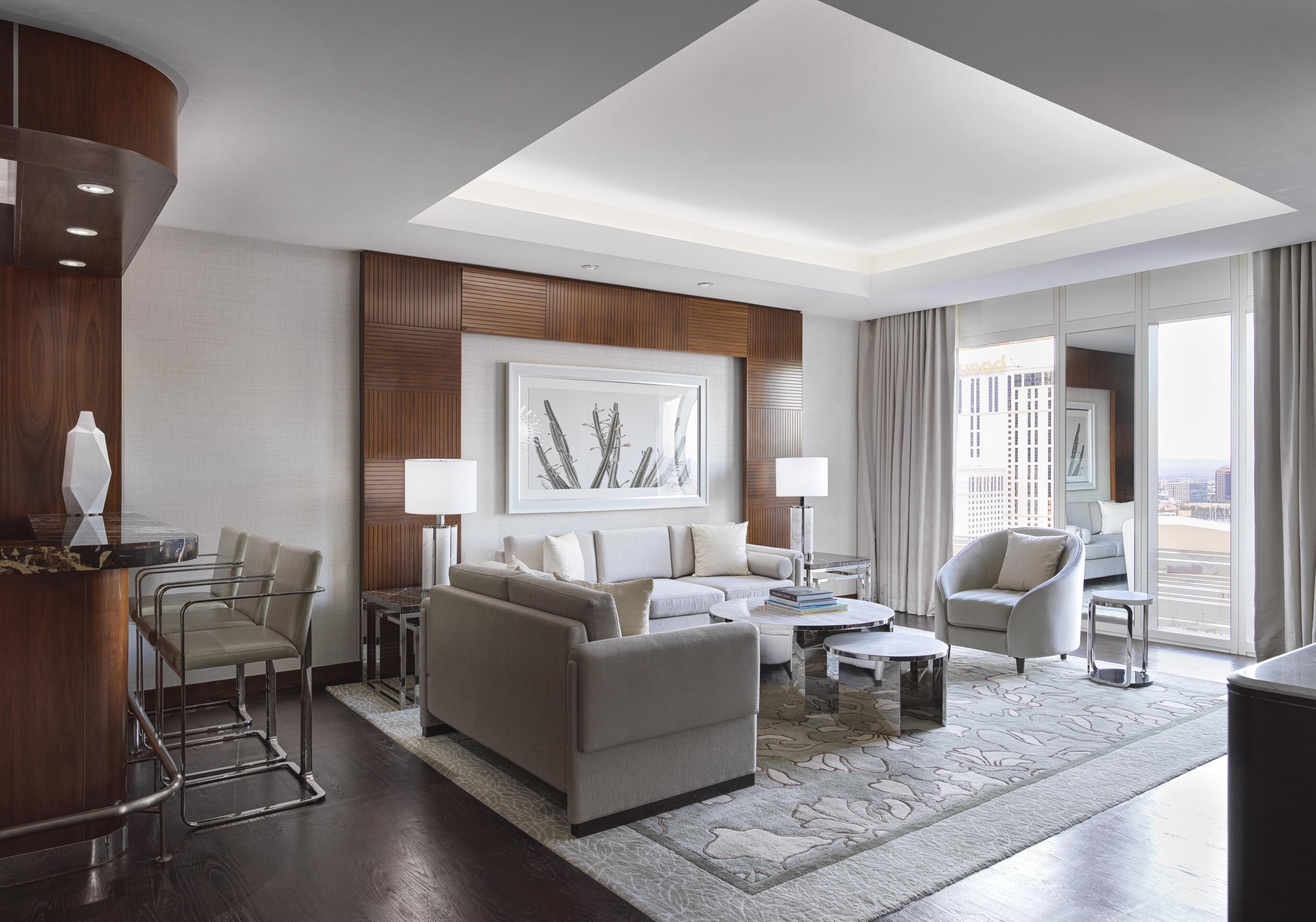 A modern hotel room with a living area featuring a sofa, armchairs, a coffee table, and a dining table. There is a large window offering a view of the city.