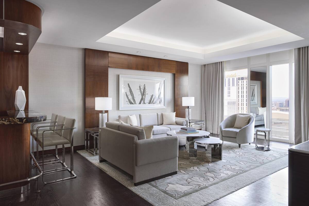 Une chambre d’hôtel moderne dotée d’un espace salon comprenant un canapé, des fauteuils, une table basse et une table à manger. Une grande fenêtre offre une vue sur la ville.
