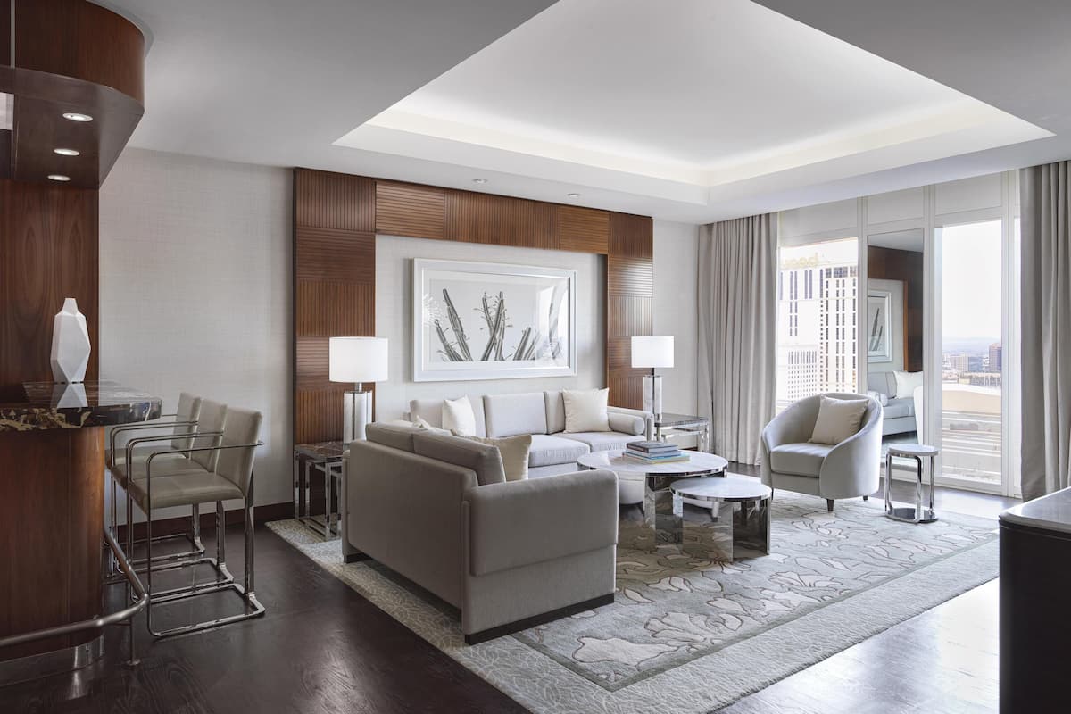 A modern hotel room with a living area featuring a sofa, armchairs, a coffee table, and a dining table. There is a large window offering a view of the city.
