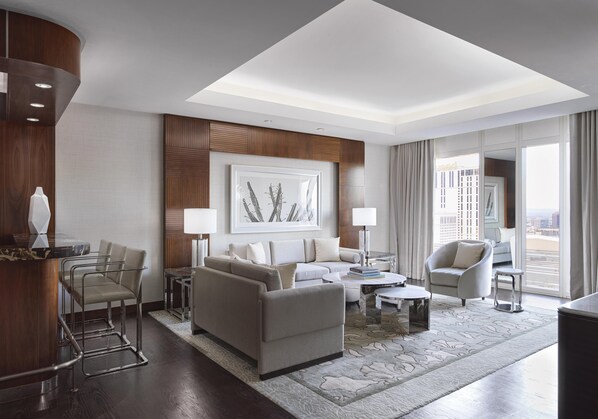 Une chambre d’hôtel moderne dotée d’un espace salon comprenant un canapé, des fauteuils, une table basse et une table à manger. Une grande fenêtre offre une vue sur la ville.