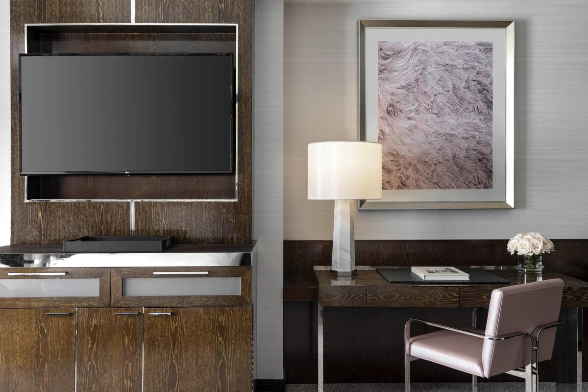 Une chambre d’hôtel moderne dotée d’un grand téléviseur à écran plat fixé sur un panneau en bois, d’un bureau avec une lampe, d’une chaise et d’un tableau abstrait encadré accroché au mur.