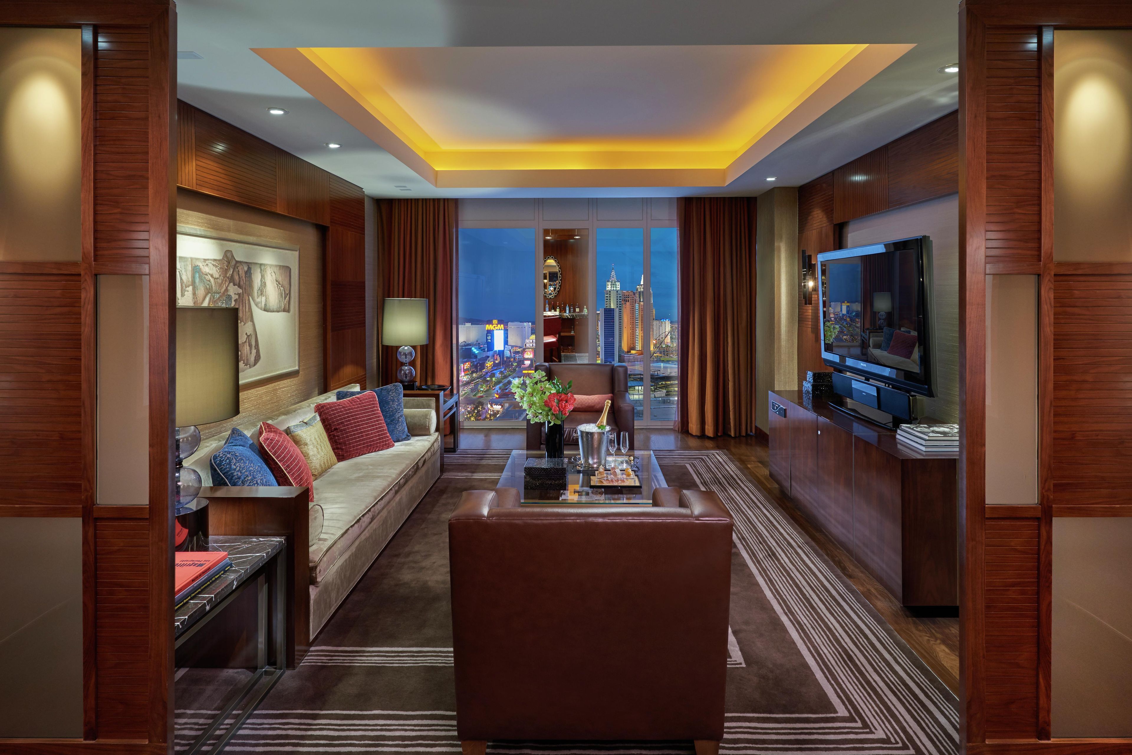 A modern hotel room with a city view, a large flat-screen TV, a sofa, a coffee table, and a dining area.
