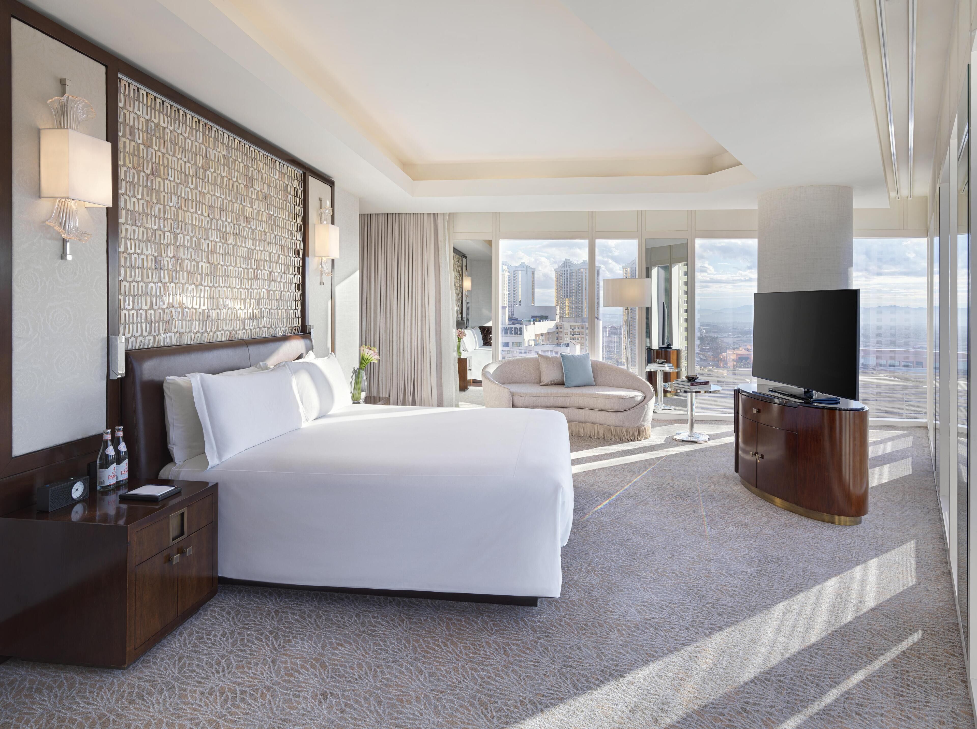 A modern hotel room with a large bed, a sofa, a TV, and a city view.