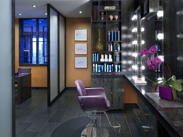 Un salón moderno con una recepción, sillas moradas, un tocador con espejos y una estantería con productos para el cabello.