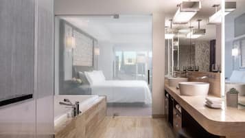 A modern hotel bathroom with a glass shower enclosure, a large bed, a vanity with a sink, and a view of the city.