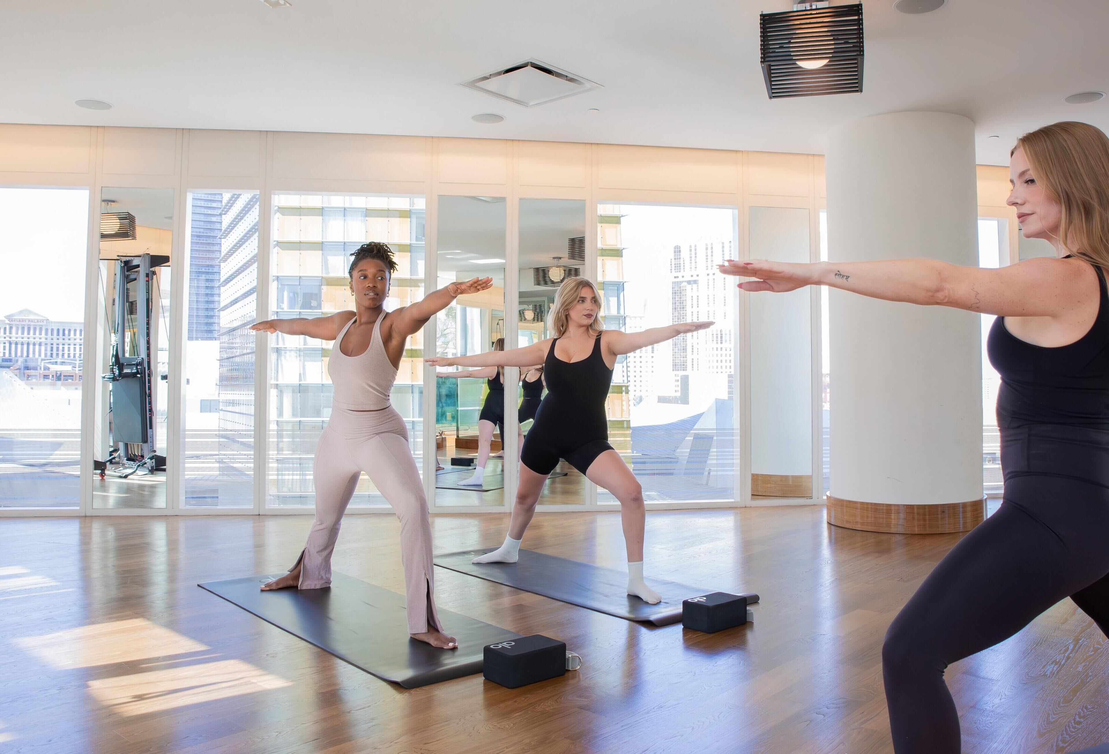 A group of people in a spacious room with large windows, wooden floors, and exercise mats.